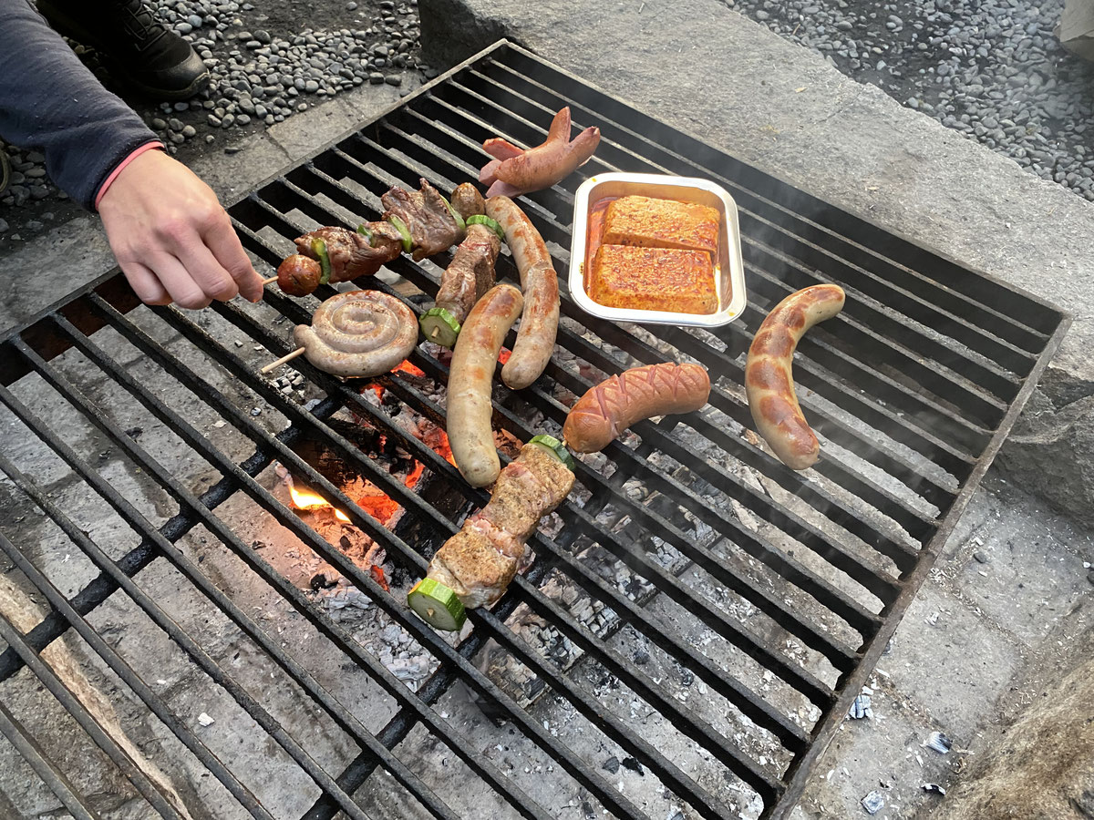 Würste und Spiesse auf einem Grillrost über offenem Feuer an einer Feuerstelle.